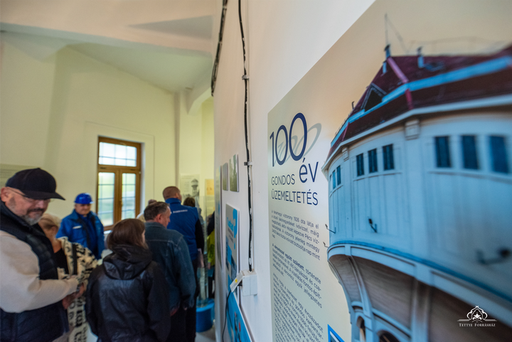 A fehérhegyi víztorony 100. születésnapja alkalmából megrendezett nyíltnapjára érkező vendégek nézelődnek a torony és a vízmű történelmét bemutató kiállításon. A kép jobb oldalán, a kiállítás egyik tablója látható, rajta a 100 éves víztrony.