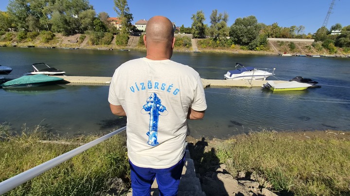 A Tettye Forrásház egyik versenyzője fehér pólóban egy vízparton áll egy lépcsőn, egy korlát mellett. A fehér póló hátán a csapatuk logója, egy kék tűzcsap és a csapat neve (Vízőrség) látható.