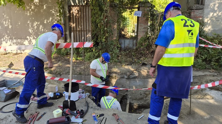 A Tettye Forrásház csapata védősisakban és láthatósági hevederben dolgozik egy elkerített munkagödör körül. Egyikük a gödörben, ketten pedig a felszínen végzik a szerelési feladatokat. A gödör mellett a versenyt felügyelő bíró védősisakban és láthatósági mellényben figyeli a munkájukat. A munkaterületen szerszámok és különböző csővezetékek, szerelvények láthatók.